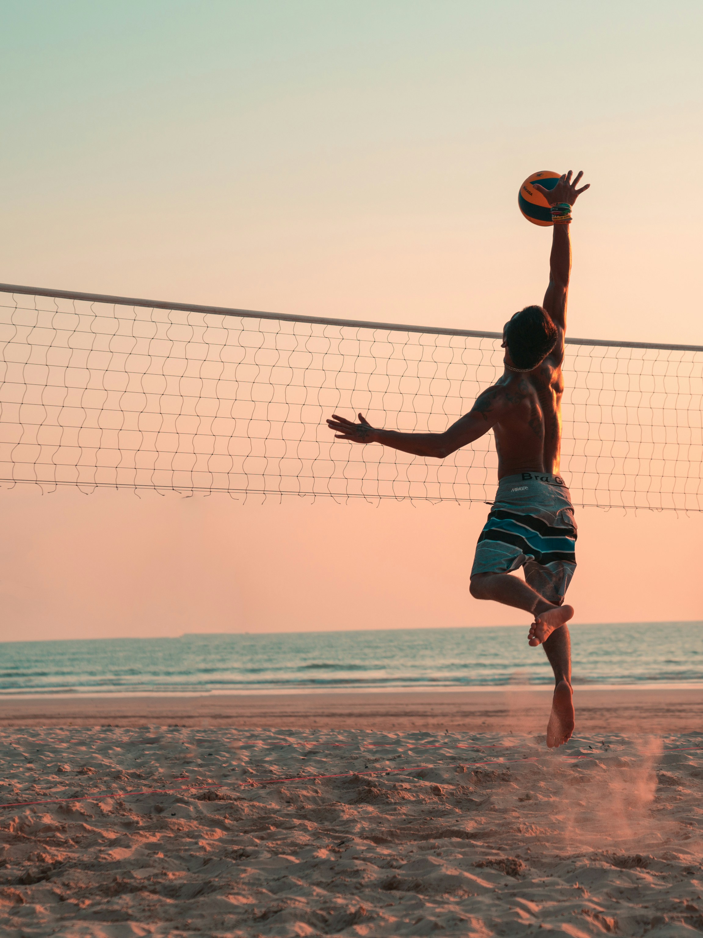 A man seen from the back jumping and hitting a volleyball over a net on the beach during the sunset.